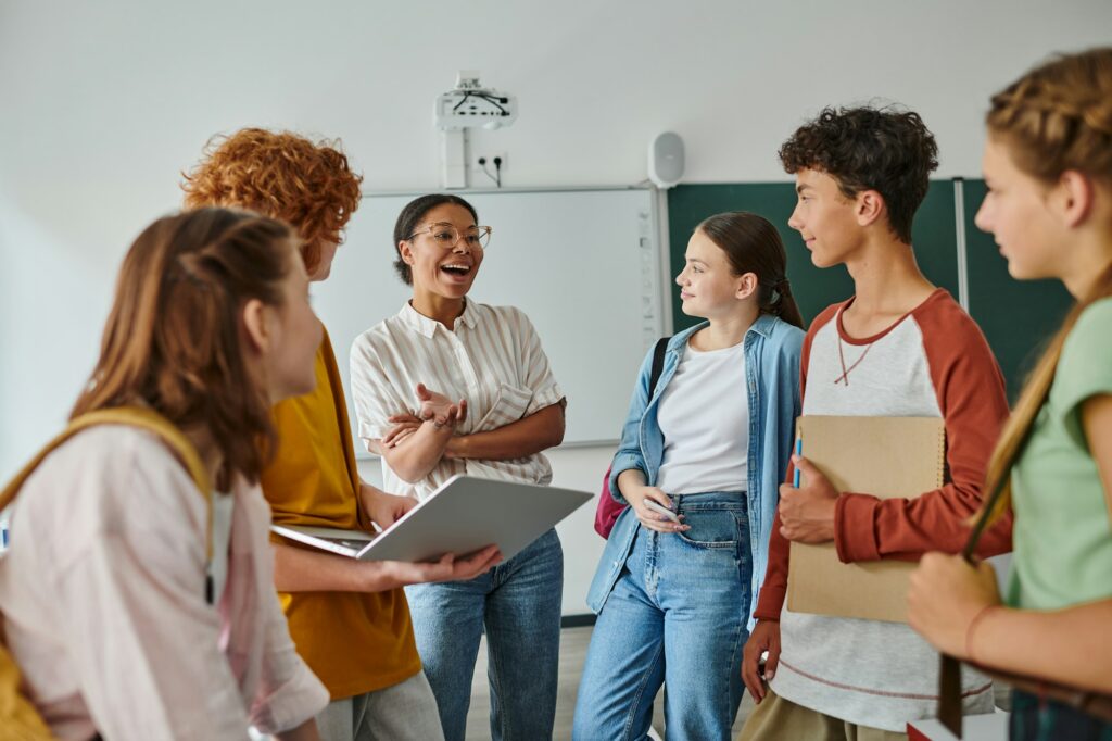 happy african american teacher talking to teenage students in classroom, back to school concept